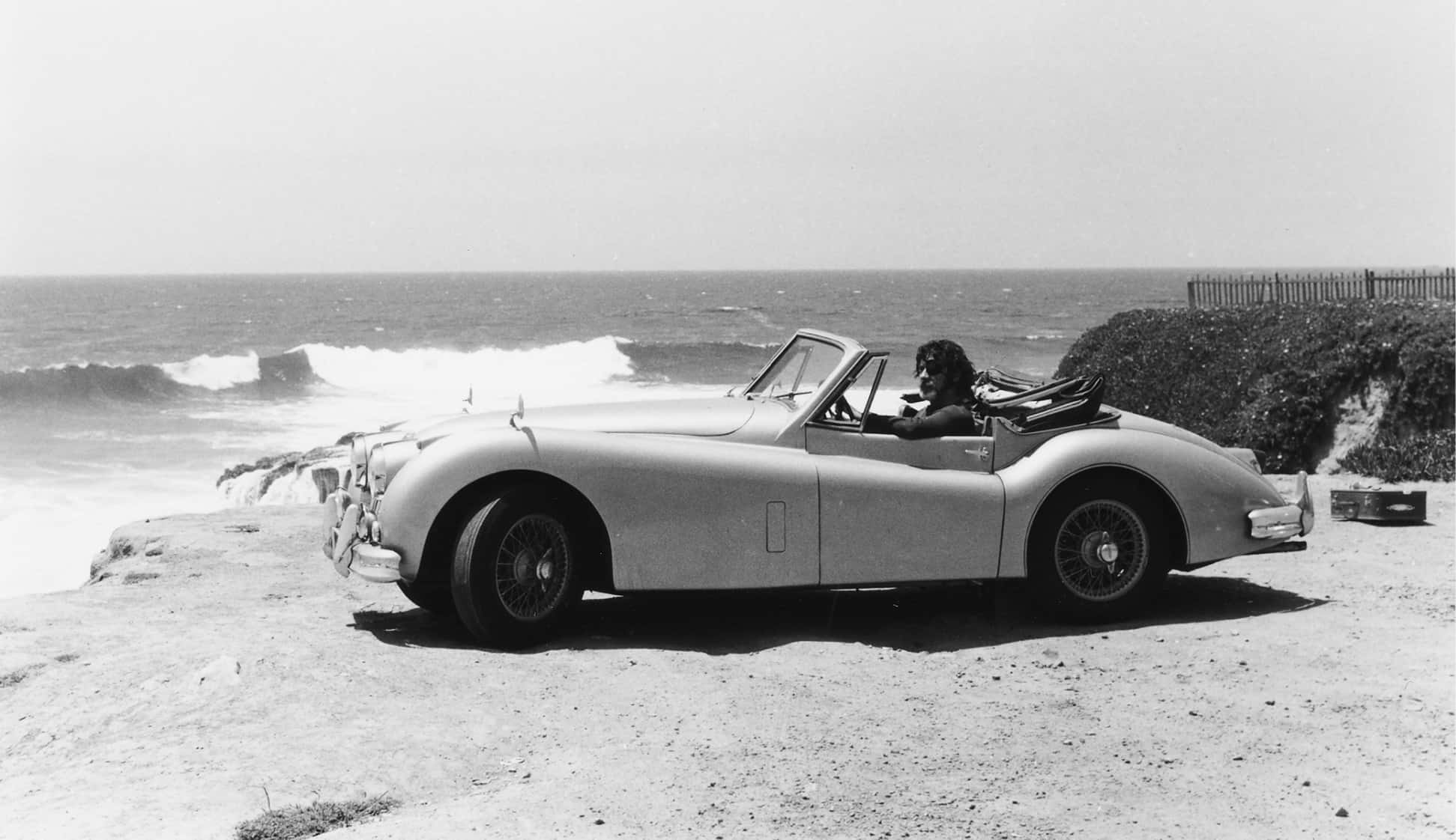A black and white photo of a classic convertible car parked by the seaside, with a person in the driver's seat enjoying the ocean view, conveying a sense of vintage leisure.