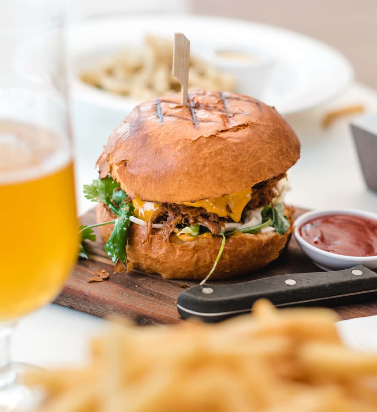 A gourmet burger with a skewer on a wooden board, served with fries, ketchup, and a glass of beer in the background, suggesting a casual dining experience.